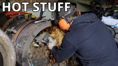 A man leans into the end of a large iron cylinder, using an angle-grinder to cut away at small metal tubes inside.